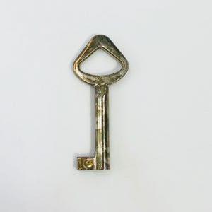 Puede incluir: Una llave metálica con un arco triangular y un vástago cilíndrico. La llave presenta un acabado plateado desgastado y deslustrado con signos visibles de uso y oxidación. El paletón es rectangular y tiene una pequeña hendidura circular.