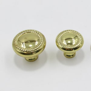 Pode incluir: Close-up de dois puxadores de armário dourados. Os puxadores têm um topo redondo e plano com uma borda em forma de corda. A base dos puxadores é cilíndrica. Os puxadores são provavelmente para móveis ou armários.