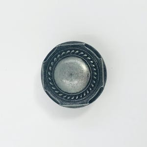 May include: A circular metal cabinet knob with a dark, weathered silver finish. The knob features a smooth, raised center dome surrounded by a textured, rope-like border and a wide, flat outer rim with subtle indentations along the edge.