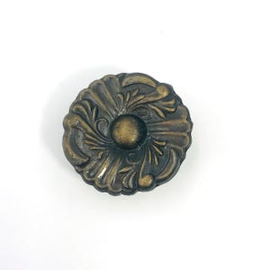 Puede incluir: Un pomo de mueble redondo de metal con acabado en latón antiguo. La superficie presenta un patrón floral simétrico en relieve con segmentos curvos en forma de pétalos que irradian desde un centro circular liso, creando un aspecto texturizado.