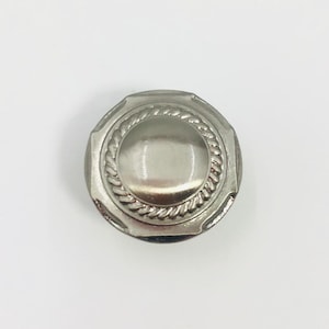 May include: A circular silver-toned metal button featuring a smooth, domed center surrounded by a raised, twisted rope-style border. The outer edge of the button has a flat, octagonal-shaped rim with a polished, reflective metallic finish.