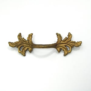 May include: A gold-colored, ornate drawer pull with a decorative leaf design. The handle is a straight bar with elaborate, symmetrical floral embellishments on each end. The hardware is likely made of metal and is designed for furniture.