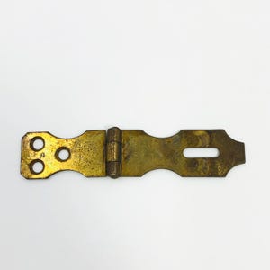 May include: A vintage brass hinge and latch set. The latch has a rectangular shape with a slotted opening and a decorative edge. The hinge side has two circular holes and two smaller holes. The metal has a tarnished, aged appearance.