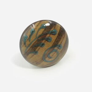 May include: A round ceramic cabinet knob with a glossy brown finish featuring vertical streaks and a hand-painted teal blue pattern of stylized leaves and a swirl. The surface is smooth and reflective, showing a distinct contrast between the brown base and the teal decorative elements.