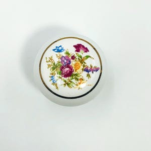 Może przedstawiać: Białe ceramiczne pokrętło do szafki z kwiatowym wzorem. Pokrętło ma złoty rant i bukiet kwiatów w odcieniach fioletu, różu, niebieskiego i żółtego. Kwiatowy wzór jest wyśrodkowany na powierzchni pokrętła.