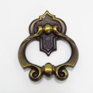 May include: A gold and brown ornate door pull with a decorative ring handle. The pull has a detailed design with scrolls and a central knob.