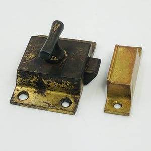 May include: Antique brass door latch with a dark handle and a matching strike plate. The latch is square with rounded corners and two screw holes. The strike plate is rectangular with one screw hole.