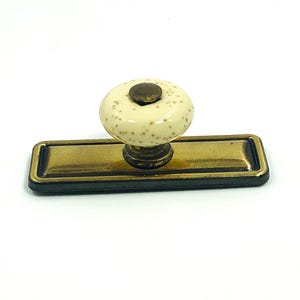 May include: A decorative cabinet pull with a cream-colored, speckled knob and a rectangular brass-toned base. The knob has a dark brown center. The base has a black border. This hardware is suitable for drawers and cabinets.