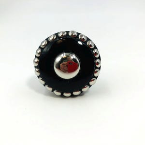 May include: A circular black cabinet knob featuring a polished silver-tone center dome and a border of small, rounded silver-tone studs around the outer edge. The reflective surface of the central dome shows a distorted red object reflection.