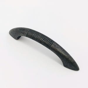 May include: A curved cabinet handle with a dark, weathered metallic finish. The centre section of the handle features a series of parallel, recessed horizontal grooves, while the ends taper to smooth, rounded points for a sleek, functional design.