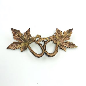 May include: A bronze-coloured drawer pull with a leaf design. It features two large maple leaves on either side of two curved handles. The leaves are detailed with veins and have a gold and brown gradient.