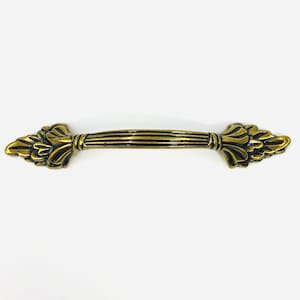 May include: A horizontal drawer pull handle finished in an antique brass tone. The central grip features a series of parallel vertical ridges, while each end is adorned with a decorative, symmetrical leaf-like motif with dark recessed accents.