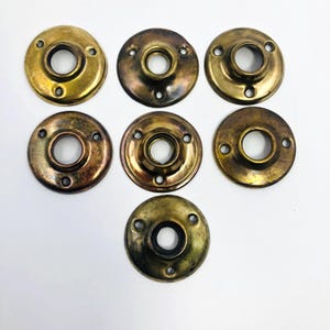 Pode incluir: Sete rosetas de porta antigas em tons de latão sobre fundo branco. Cada uma tem um orifício central e três orifícios para parafusos. O acabamento metálico varia de dourado a bronze, mostrando sinais de desgaste.