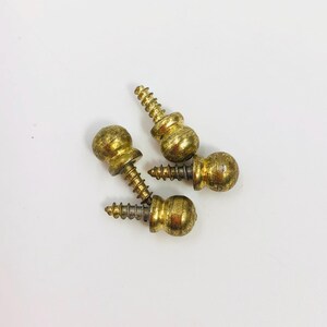 Può includere: Tre pomelli a vite in metallo color ottone, ciascuno caratterizzato da una testa sferica con parte superiore liscia e arrotondata e un gambo filettato in metallo che si estende dalla base, disposti in gruppo su una superficie chiara.
