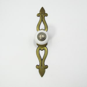 May include: A decorative brass cabinet pull with a white ceramic knob and a silver-toned center. The pull has an ornate, elongated design with a pointed top and bottom, and heart-shaped cutouts. The hardware is designed for furniture.