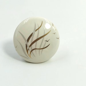Op de afbeelding: Een ronde, crèmekleurige keramische knop met een op de natuur geïnspireerd ontwerp. De knop heeft goud en grijs gestileerd gras en vliegende vogels. Geschikt voor kasten, lades of meubels.