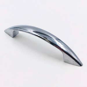 May include: A chrome-finished cabinet pull handle. The handle has a curved, arched design with rectangular mounting bases on each end. The surface reflects light, highlighting its polished appearance. This hardware is suitable for cabinets and drawers.