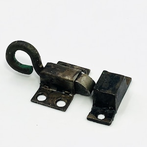 May include: An antique metal latch with a hook and eye closure. The latch is made of dark metal with a weathered appearance. The hook is curved and has a green patina. The latch is designed to secure a door or cabinet.