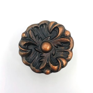 Puede incluir: Un pomo de mueble redondo de metal con acabado en cobre oscuro. El pomo presenta un patrón floral simétrico en relieve con una cúpula circular central rodeada de segmentos curvos en forma de pétalos y pequeñas cuentas redondeadas.