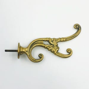 May include: Ornate brass wall hook with a decorative, scroll-like design. The hook features a screw for mounting and a detailed floral or foliate motif. The brass has a warm, aged patina, suggesting a vintage or antique aesthetic.