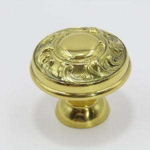 May include: A gold-colored door knob with an ornate, swirling design. The knob has a rounded top with a central, raised circle and sits on a short, pedestal base. The metal has a polished, reflective surface.