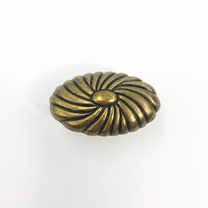 May include: An oval-shaped metal cabinet knob with an antique brass finish. The surface features a raised, swirling radial pattern that radiates from a smooth, solid center point, creating a textured, dimensional appearance across the entire piece.