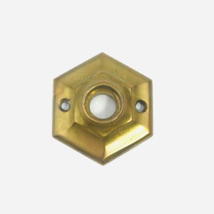 Puede incluir: Una placa de puerta hexagonal de latón con un acabado metálico pulido. La placa presenta una abertura circular central para un eje y dos orificios circulares más pequeños a la izquierda y a la derecha para los tornillos de montaje.