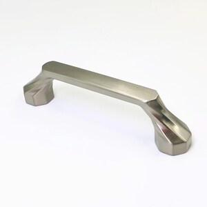 May include: A brushed nickel cabinet handle with a smooth, flat top bar and faceted, geometric mounting bases on each end. The metal hardware features a metallic silver finish with a subtle sheen and a clean, angular design throughout the piece.