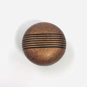 May include: A round cabinet knob with a textured, aged copper finish. The surface features five horizontal, recessed black lines running across the center of the sphere, creating a distinct contrast against the mottled brown metallic surface.