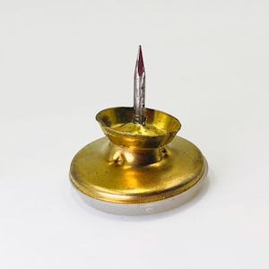 May include: A gold-toned metal thumbtack featuring a wide, circular base and a central, sharp, silver-colored steel needle point. The metallic surface shows a slightly weathered texture with a reflective finish, standing upright on its flat bottom.