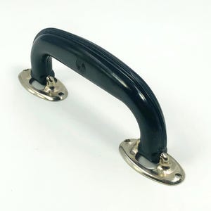 Puede incluir: Un asa negra y arqueada con un agarre acanalado, fijada a dos placas de montaje ovaladas de color plateado. El asa es probablemente para una maleta vintage o un artículo similar. Las placas tienen pequeños detalles decorativos.