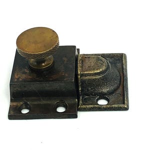 May include: A vintage bronze window latch with a round knob and a rectangular base. The latch has a tarnished appearance, suggesting age and use. The design includes a catch and a knob for easy opening and closing.