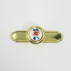 May include: A gold-toned metal cabinet pull featuring a rectangular backplate with rounded ends and a central circular knob. The knob has a white ceramic insert decorated with a small, colourful floral pattern of pink and blue flowers.