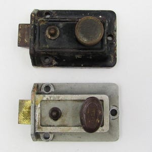 Könnte beinhalten: Zwei antike Türriegel, einer schwarz und einer silberfarben, mit runden Knöpfen. Beide Riegel sind rechteckig und aus Metall.