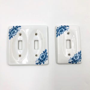 Könnte beinhalten: Zwei weiße Lichtschalterabdeckungen aus Keramik mit blauem Blumenmuster in den Ecken. Eine Abdeckung ist rechteckig für einen einzelnen Schalter, die andere ist breiter und für zwei Kippschalter mit entsprechenden Schraublöchern ausgelegt.