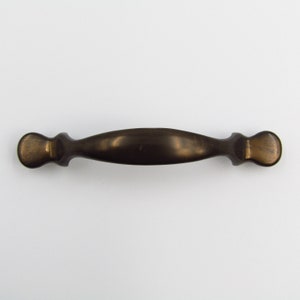 Könnte beinhalten: Ein bronzefarbener Schrankgriff mit einer gebogenen, länglichen Mitte und abgerundeten Enden. Die Hardware hat eine Vintage-Ästhetik und eignet sich für Schubladen oder Schranktüren. Der Griff ist vor einem weißen Hintergrund platziert.