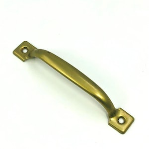 May include: A brass-colored cabinet pull handle with a curved bar and square mounting plates. The handle is approximately 4 inches long and has a smooth, slightly aged finish. The mounting plates have a single hole for screws.