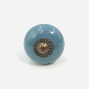 May include: A round, smooth, dusty blue ceramic cabinet knob with a glossy finish. The center features a decorative, dark metallic, scalloped-edge circular plate with a small, raised, rounded metal dome in the middle of the hardware piece.