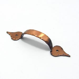 以下が含まれることがあります： 銅色のキャビネットプルで、湾曲したハンドルと装飾的な端部を備えています。ハンドルはハンマーで叩かれたような質感で、リベットで端部に固定されています。端部は葉のような形をしています。