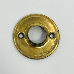 May include: A vintage brass door knob backplate with a circular design. The plate has a central hole and two smaller holes for screws. The brass has a worn, aged appearance with areas of discoloration, suggesting its age and use.