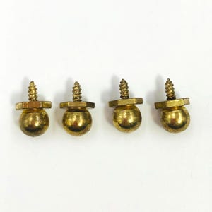 Può includere: Quattro pomelli per mobili in metallo color ottone, ciascuno caratterizzato da una testa sferica fissata a una base esagonale con una vite filettata che sporge dalla parte superiore. La ferramenta ha una finitura lucida e dorata.