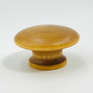 Puede incluir: Un tirador de armario de madera con forma de hongo. El tirador tiene una parte superior redondeada y una base cilíndrica. La veta de la madera es visible y el color es marrón claro natural. Adecuado para cajones y armarios.