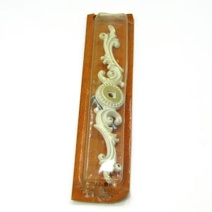 May include: A white, ornate furniture applique with a scroll design, packaged in a clear plastic blister pack on a brown cardboard backing. The applique features a central circular element and is approximately 6 inches long.