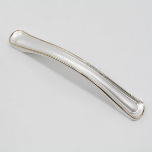May include: A silver-colored cabinet pull with a gold-colored trim. The handle is curved and has a rectangular shape with rounded ends. The hardware is designed for furniture and home decor.