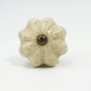 May include: A flower-shaped cabinet knob with a cream-colored ceramic surface featuring a dark brown crackle glaze finish. The center of the knob has a small, circular, metallic-toned hardware piece that is recessed into the ceramic material.