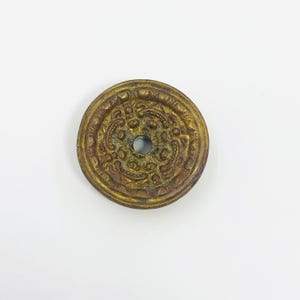 May include: A circular brass button featuring a raised, intricate relief pattern across its surface. The design includes curved lines and small dots surrounding a central hole. The metal has a weathered, aged patina with dark brown and gold tones.