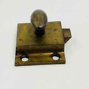 May include: An antique brass door latch with a rounded knob. The latch is rectangular with two screw holes and a small, angled piece for the latch mechanism. The brass has a weathered, aged appearance.