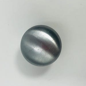 Pode incluir: Um puxador de móvel circular com acabamento em metal prateado escovado. O puxador apresenta uma superfície lisa e ligeiramente abaulada com um brilho metálico subtil e uma textura de grão fino visível na face, constituindo uma peça de ferragem limpa.