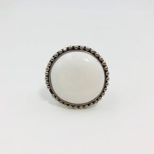 May include: A circular ring featuring a smooth, opaque white center stone or inlay. The setting is crafted from a metallic material with a beaded, textured border that encircles the white disc, creating a distinct contrast between the center and edge.