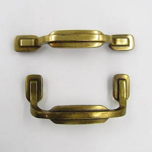 Puede incluir: Dos tiradores de armario de latón con forma rectangular y un diseño ligeramente curvo. Los tiradores tienen un acabado liso y pulido.